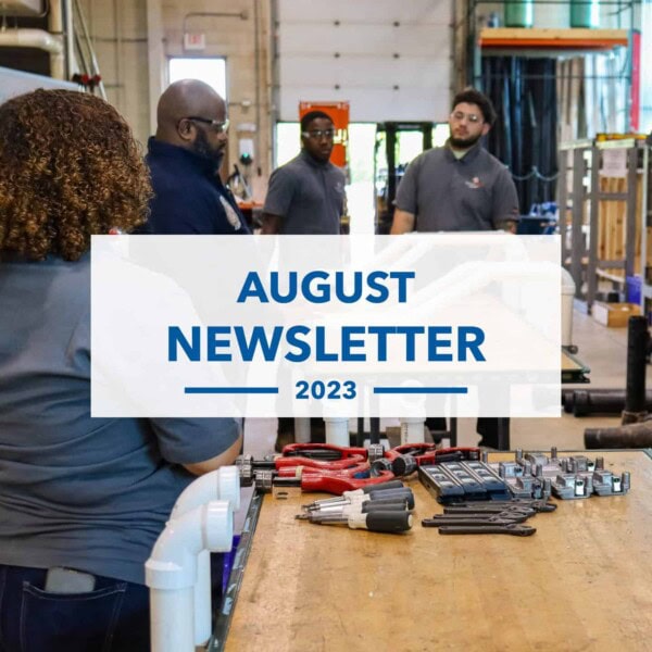 Four people in gray shirts stand and talk in a workshop with tools on a table in the foreground. Text on image reads August Newsletter 2023.