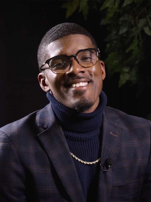 A man with short hair and glasses smiles, wearing a dark plaid blazer, navy turtleneck, gold chain, and lapel microphone. The background is dark with green leaves visible on the right.