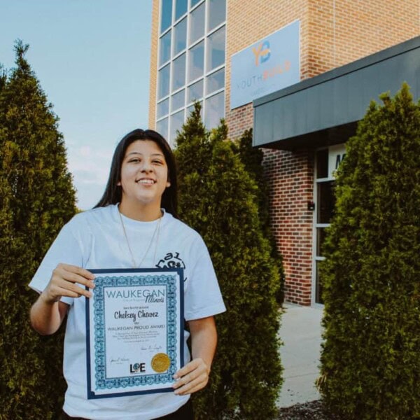 A young woman smiles while holding a framed award certificate outside a brick building with “YouthBuild” on a sign above the entrance, standing between tall green shrubs.
