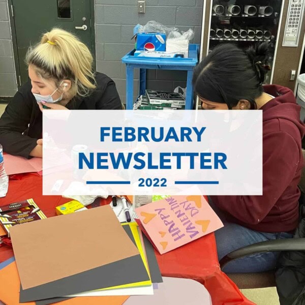 Two people sit at a table covered with colored paper and art supplies, making crafts. A sign in the center reads February Newsletter 2022. One person wears a mask and a ponytail, the other has dark hair in a bun.