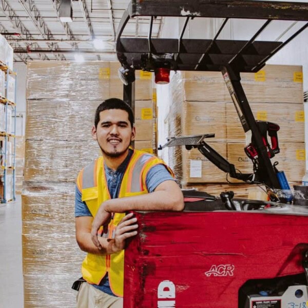 A man wearing a yellow safety vest leans on a red forklift in a warehouse, with stacked pallets of boxed goods behind him. He smiles at the camera, standing among high shelves and industrial lighting.