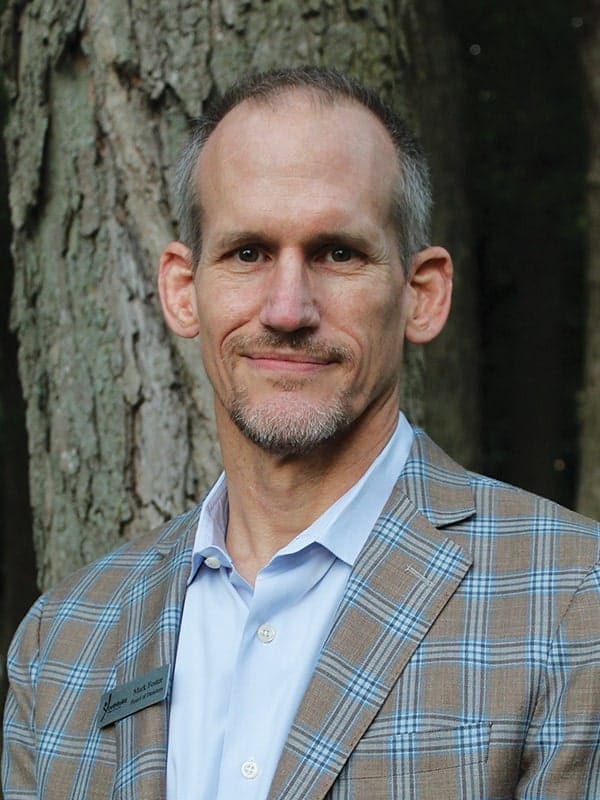A man with short hair and a trimmed beard wearing a light blue shirt and a plaid blazer stands outdoors in front of a tree, looking at the camera and smiling slightly.