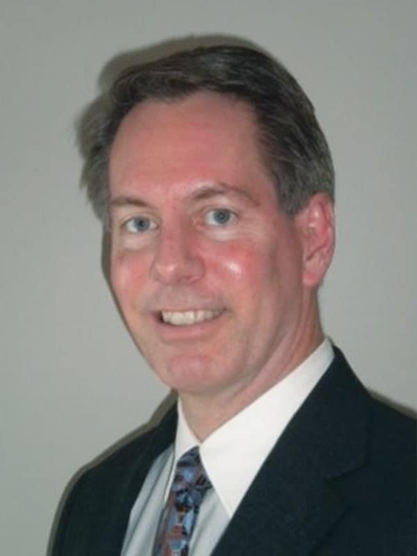 A man with short brown hair wearing a dark suit, white shirt, and patterned tie, smiling in front of a plain light-colored background.