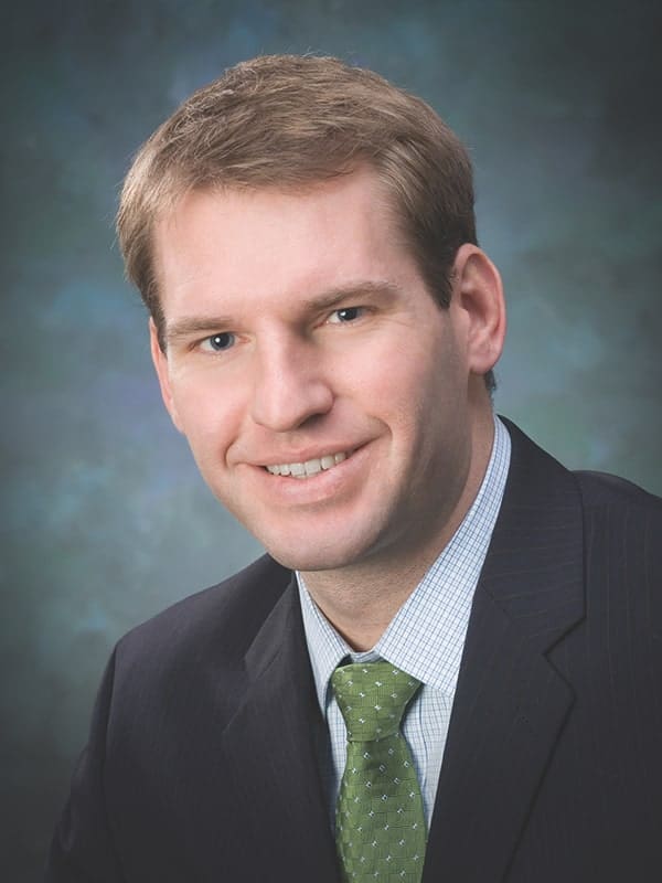 A man with short light brown hair wearing a dark suit, light blue shirt, and green tie, smiling at the camera in front of a blurred blue-gray background.