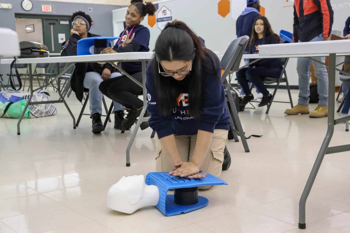 A woman kneels on the floor practicing CPR on a training mannequin while others watch in a classroom setting. Tables and chairs are arranged around the room.