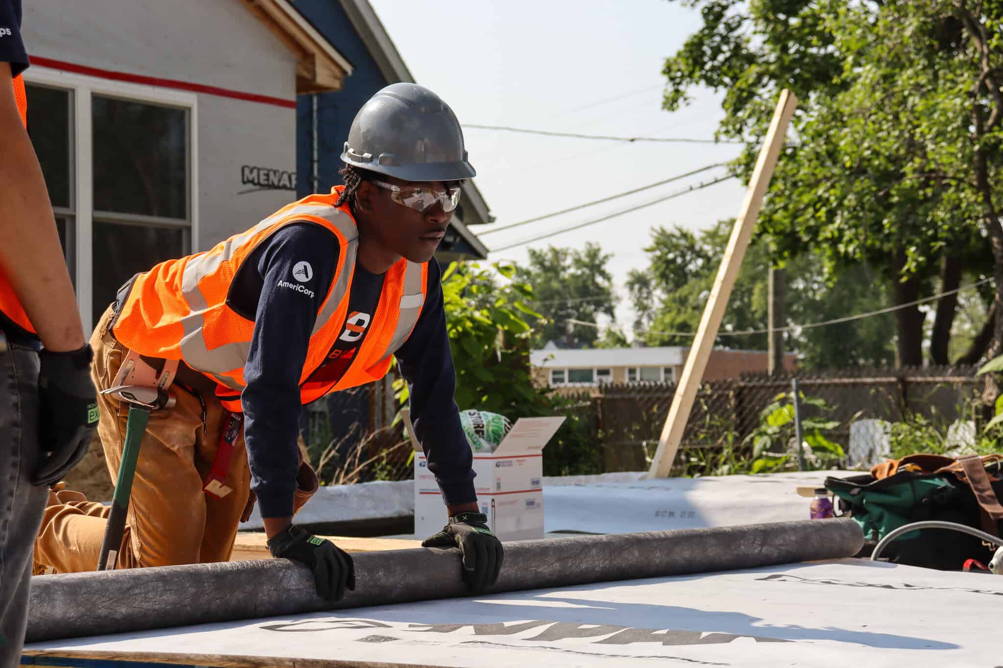 A construction worker in an orange safety vest and helmet kneels on a roof, preparing materials. Another worker stands nearby. In the background are trees, a fence, and a house.