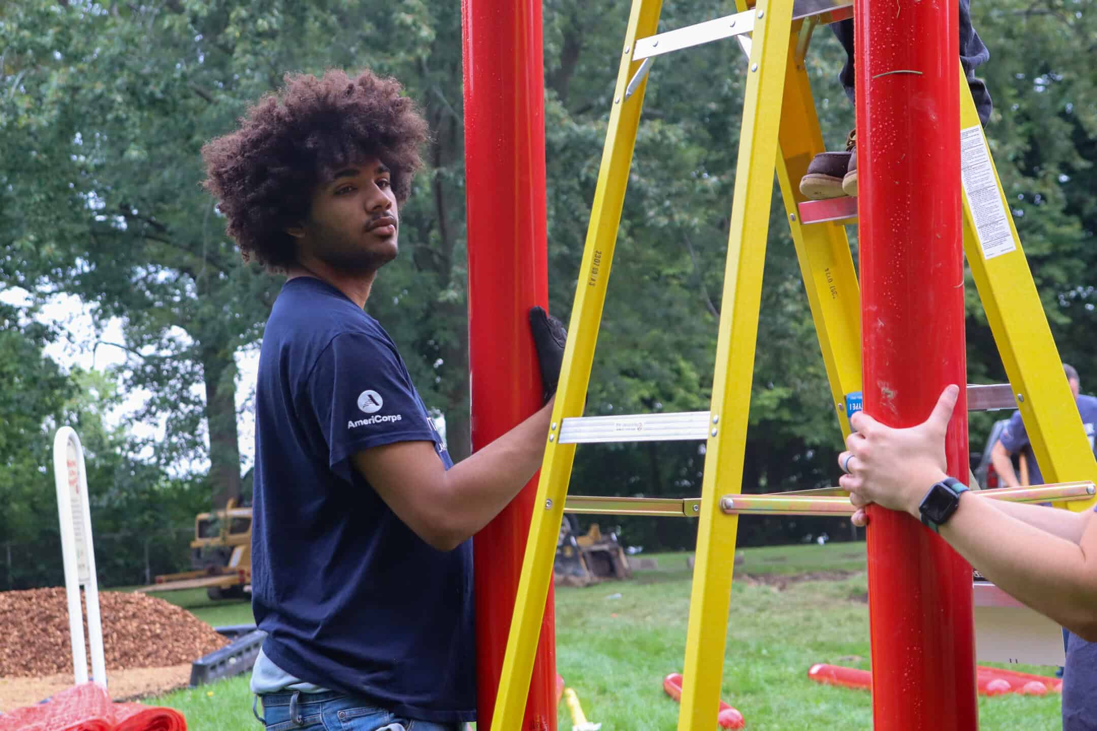 A person in a navy shirt and gloves steadies a red pole next to a yellow ladder outdoors, while another person holds the ladder. Trees and construction materials are visible in the background.