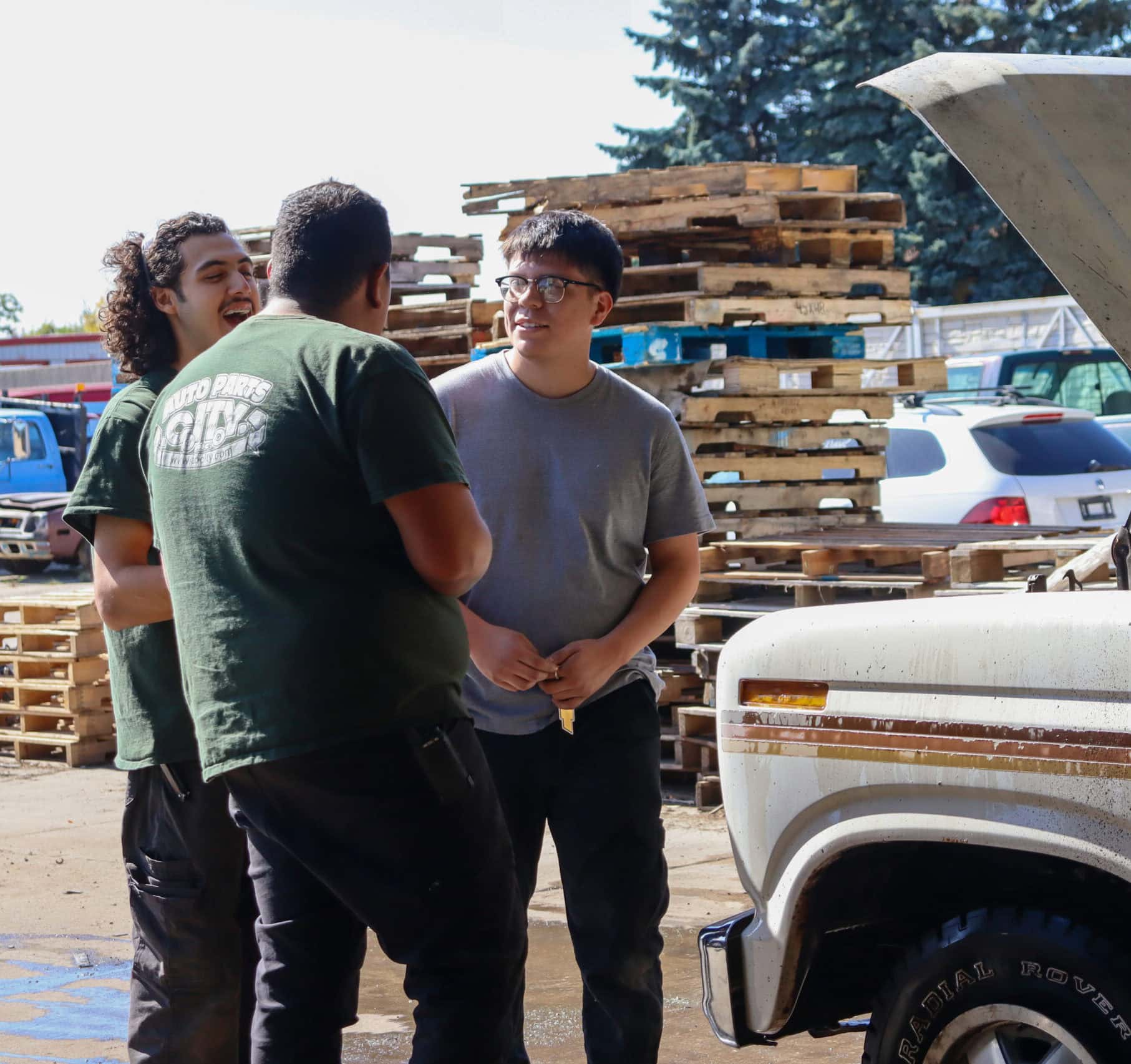 Three men stand talking near a white truck with its hood open, outdoors on a sunny day. Wooden pallets and parked cars are visible in the background.
