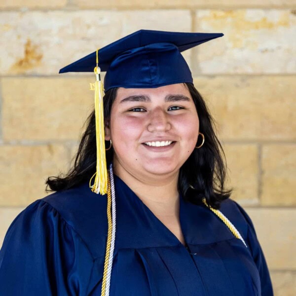 A young woman wearing a navy blue graduation cap and gown smiles at the camera, standing in front of a tan brick wall. She has long dark hair and is wearing gold hoop earrings and a tassel on her cap.