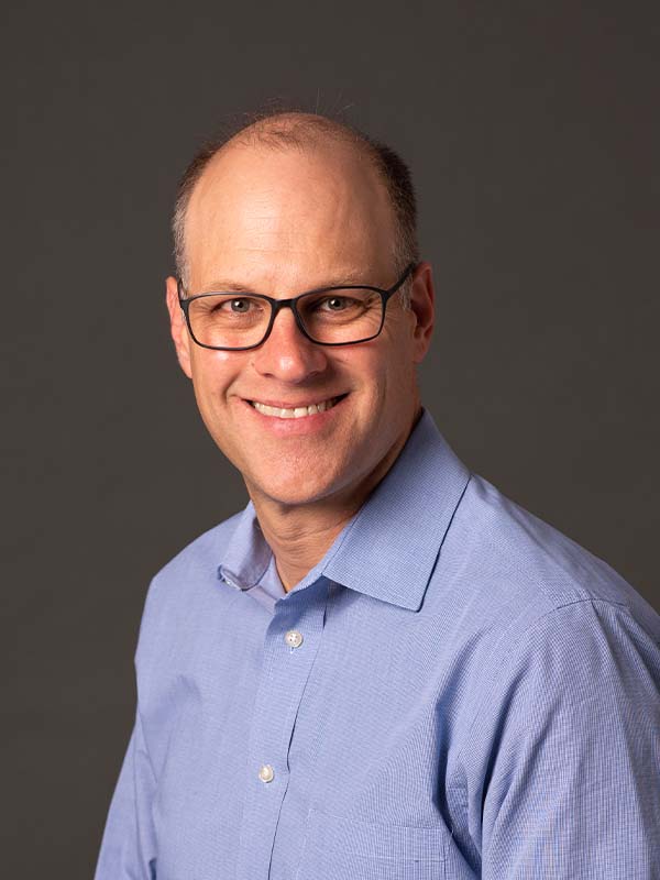 A smiling man with glasses and short hair wearing a light blue collared shirt poses in front of a plain dark background.