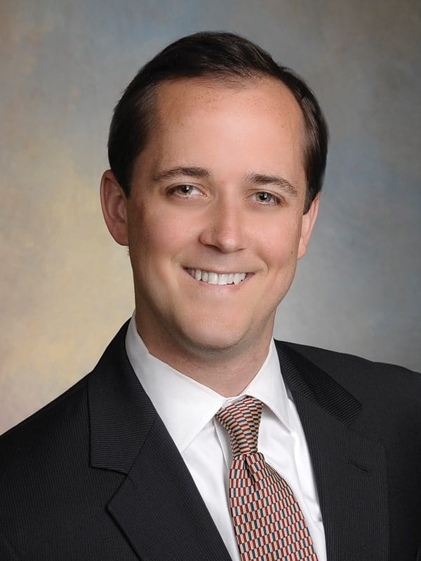A man wearing a dark suit, white shirt, and patterned tie, smiling at the camera against a neutral, softly colored studio background.