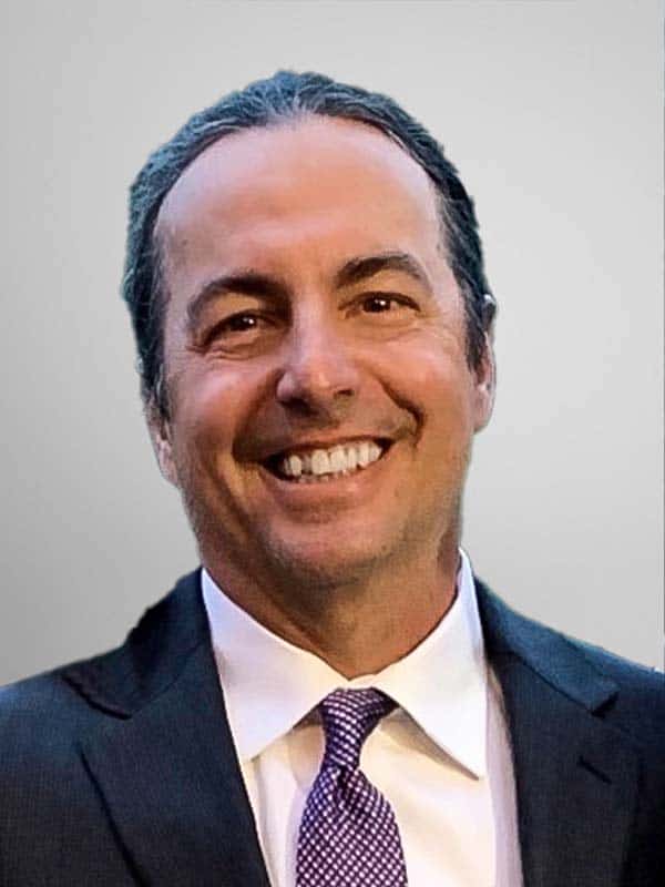 A man with dark hair, wearing a dark suit, white shirt, and purple patterned tie, smiles at the camera against a plain light gray background.