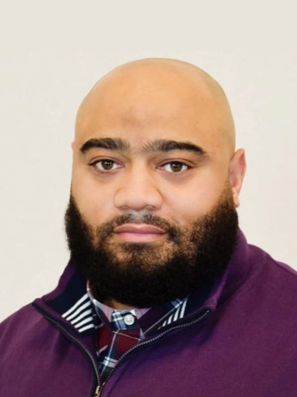 A man with a shaved head and a full, thick beard is wearing a purple zip-up jacket over a plaid shirt, posing against a plain, light background.