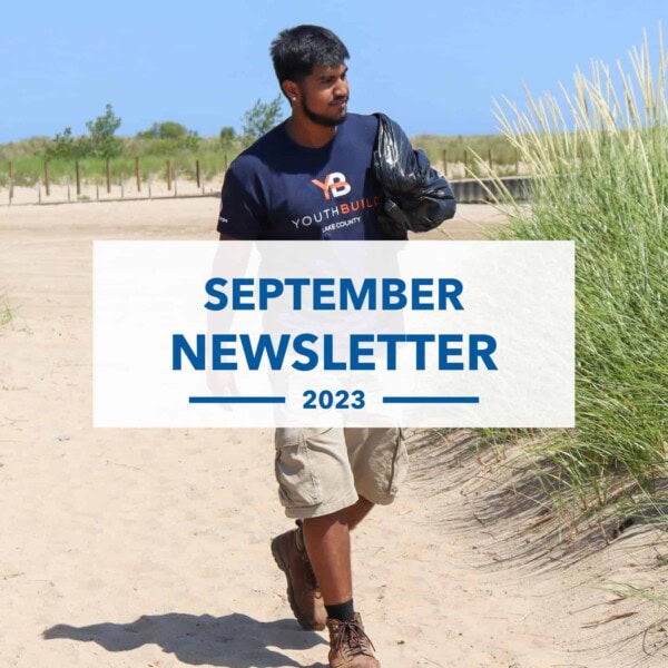A young man wearing a YouthBuild T-shirt and gloves picks up trash on a sandy beach. The text overlay reads September Newsletter 2023. Grassy dunes and blue sky are in the background.