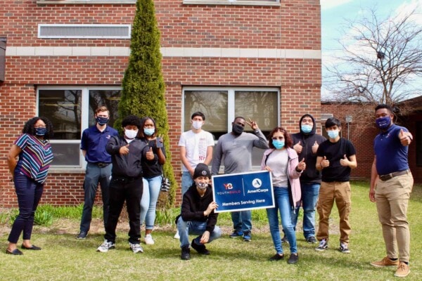 A diverse group of people wearing masks stands outside a brick building on grass, some giving thumbs up. Two people in front hold a blue AmeriCorps VISTA sign that reads “Members Serving Here.”.