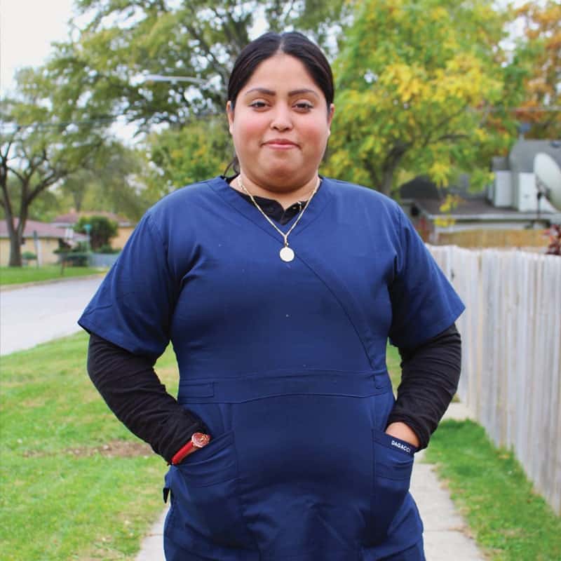 A woman wearing blue medical scrubs and a black undershirt stands outdoors on a sidewalk, with her hands in her pockets. Trees and houses are visible in the background. She is looking at the camera and smiling slightly.